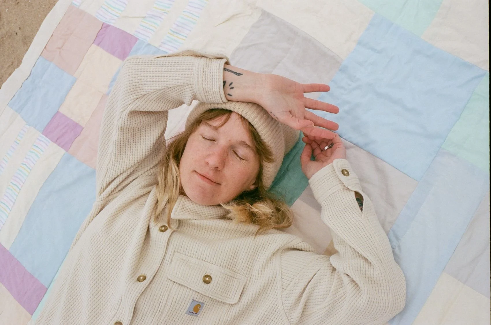 A person with blonde hair lying on a pastel patchwork quilt, wearing a beige sweater and beanie, with their eyes closed and left arm resting on their head.