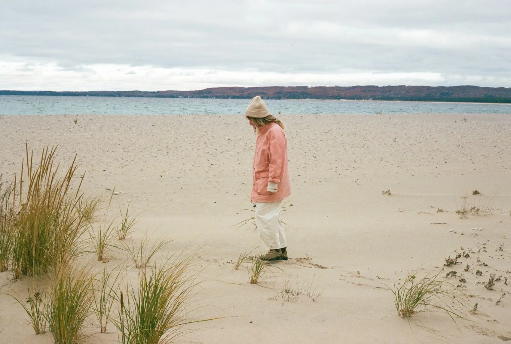 A person dressed in a pink jacket, light-colored pants, and black boots walking on a sandy beach with dune grass, near a body of water under a cloudy sky.