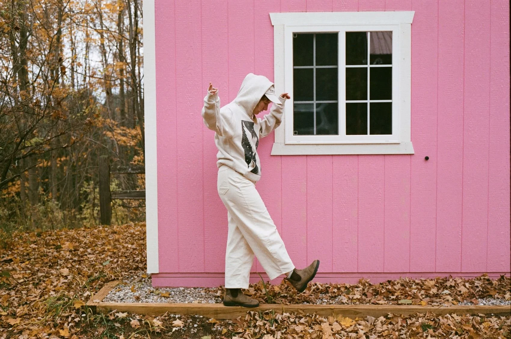 Person in a white hoodie, beige pants, and boots walking on a wooden plank path next to a pink building with a window, surrounded by fallen autumn leaves.