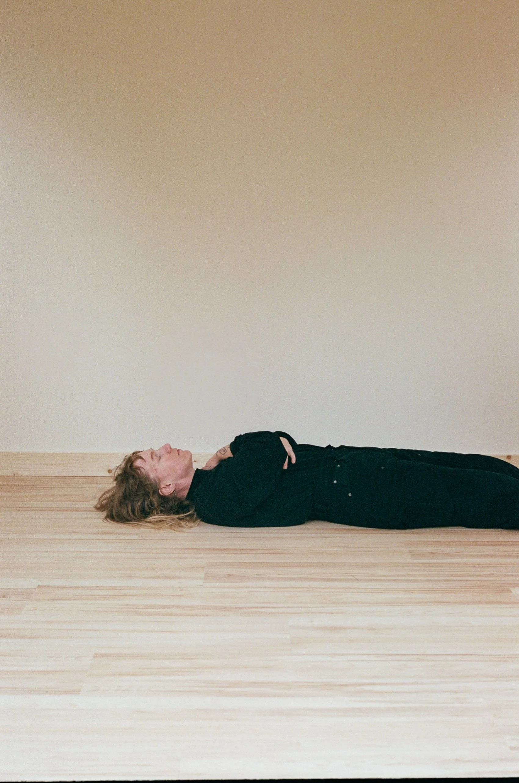 A person lying on their back on a wooden floor, with arms crossed over their chest and eyes closed.