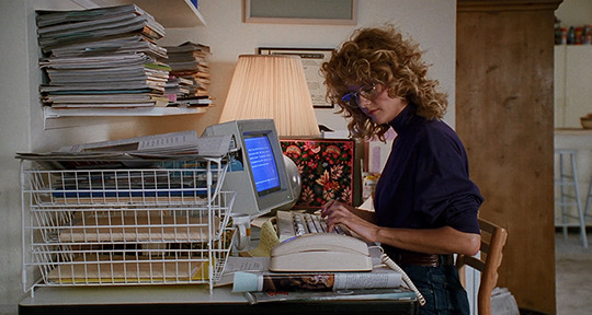A woman with curly hair wearing glasses and a purple shirt is working at a cluttered desk with a vintage computer, surrounded by stacks of paper and files, in a home office.