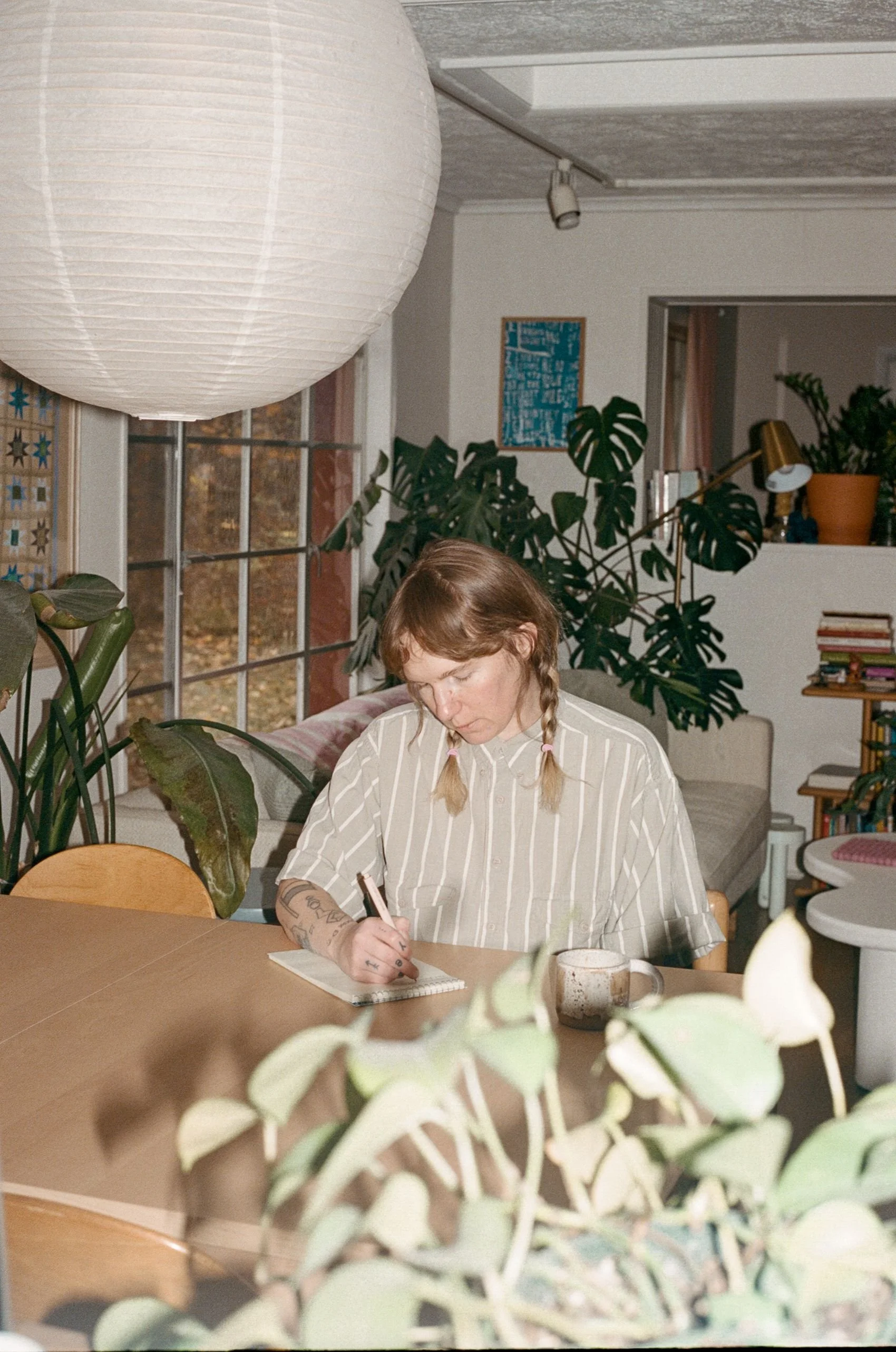 A person with blonde hair in braids, wearing a striped shirt, sitting at a wooden table, writing in a notepad with a coffee mug nearby, in a cozy, plant-filled room.
