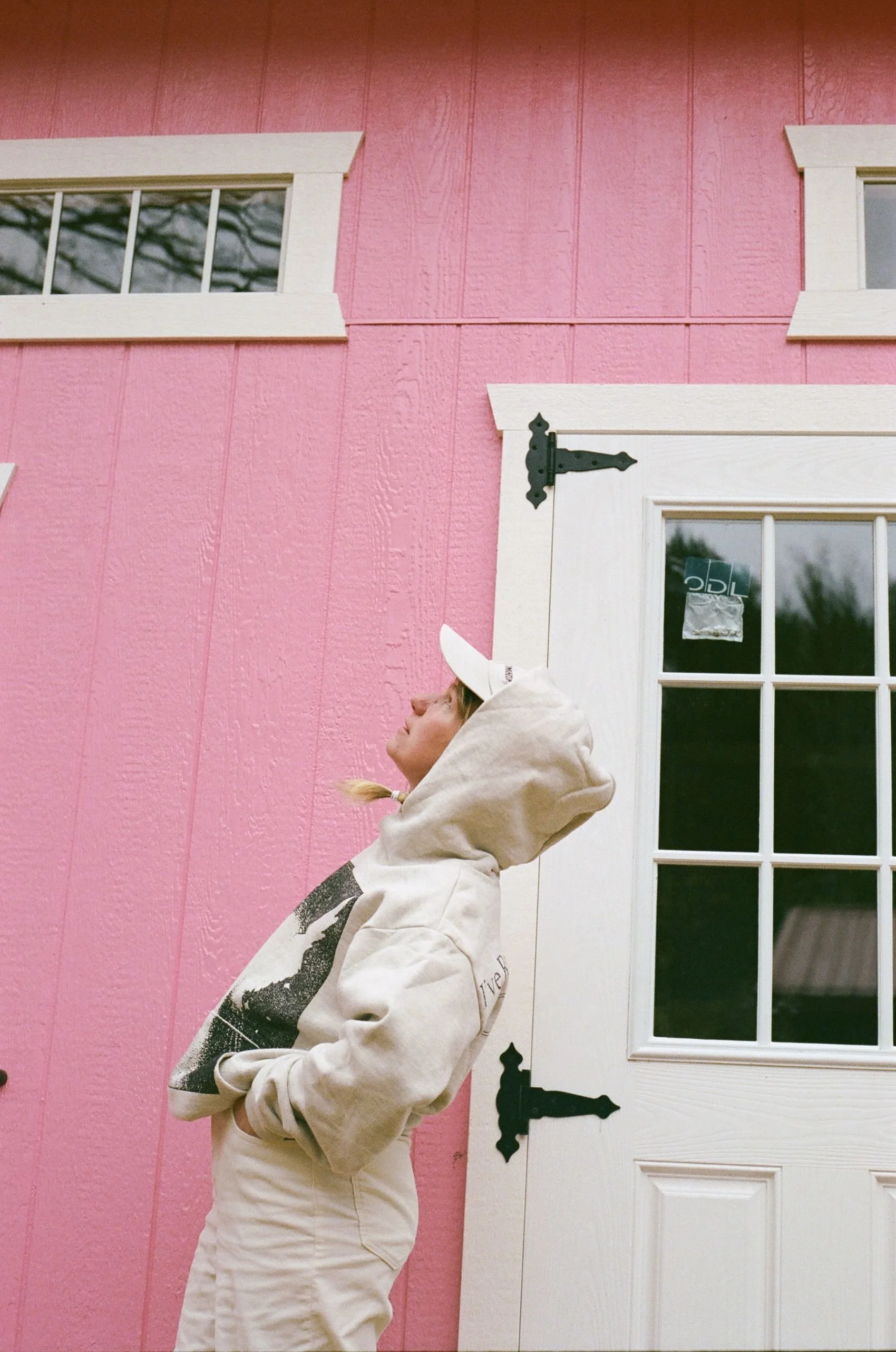 A person wearing a beige hoodie, beige pants, and a white cap stands outdoors near a pink building with white trim and black hinges, looking up.
