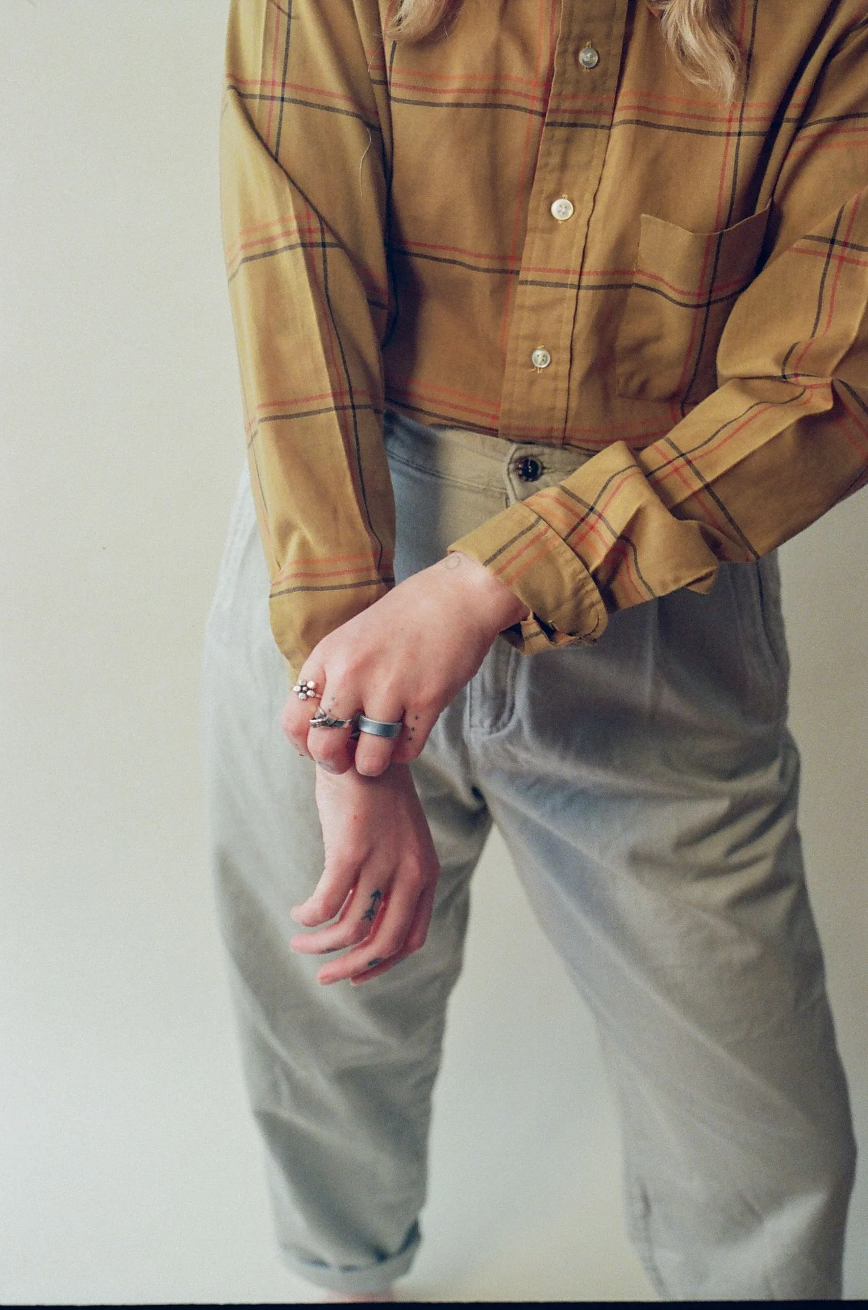 Person wearing a tan plaid button-up shirt and beige pants, with rings on fingers and visible tattoos on hands.