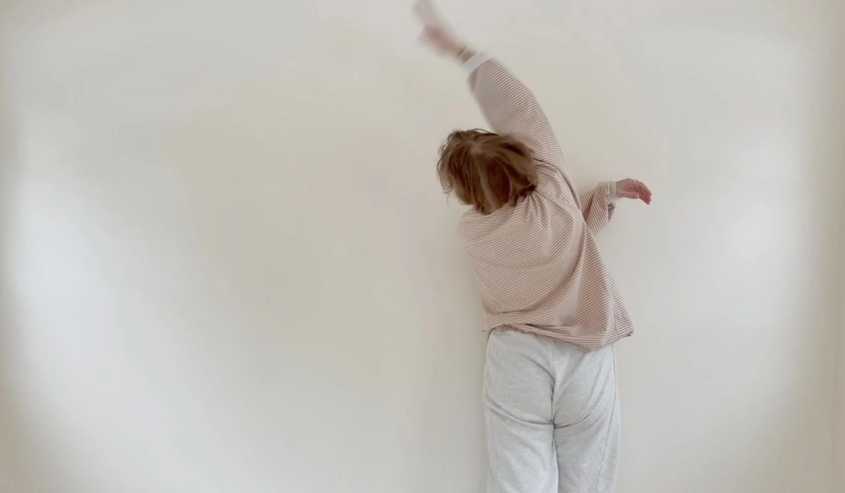 Person with short hair, wearing a beige striped top and white pants, reaching upward with one arm while standing against a plain white wall.