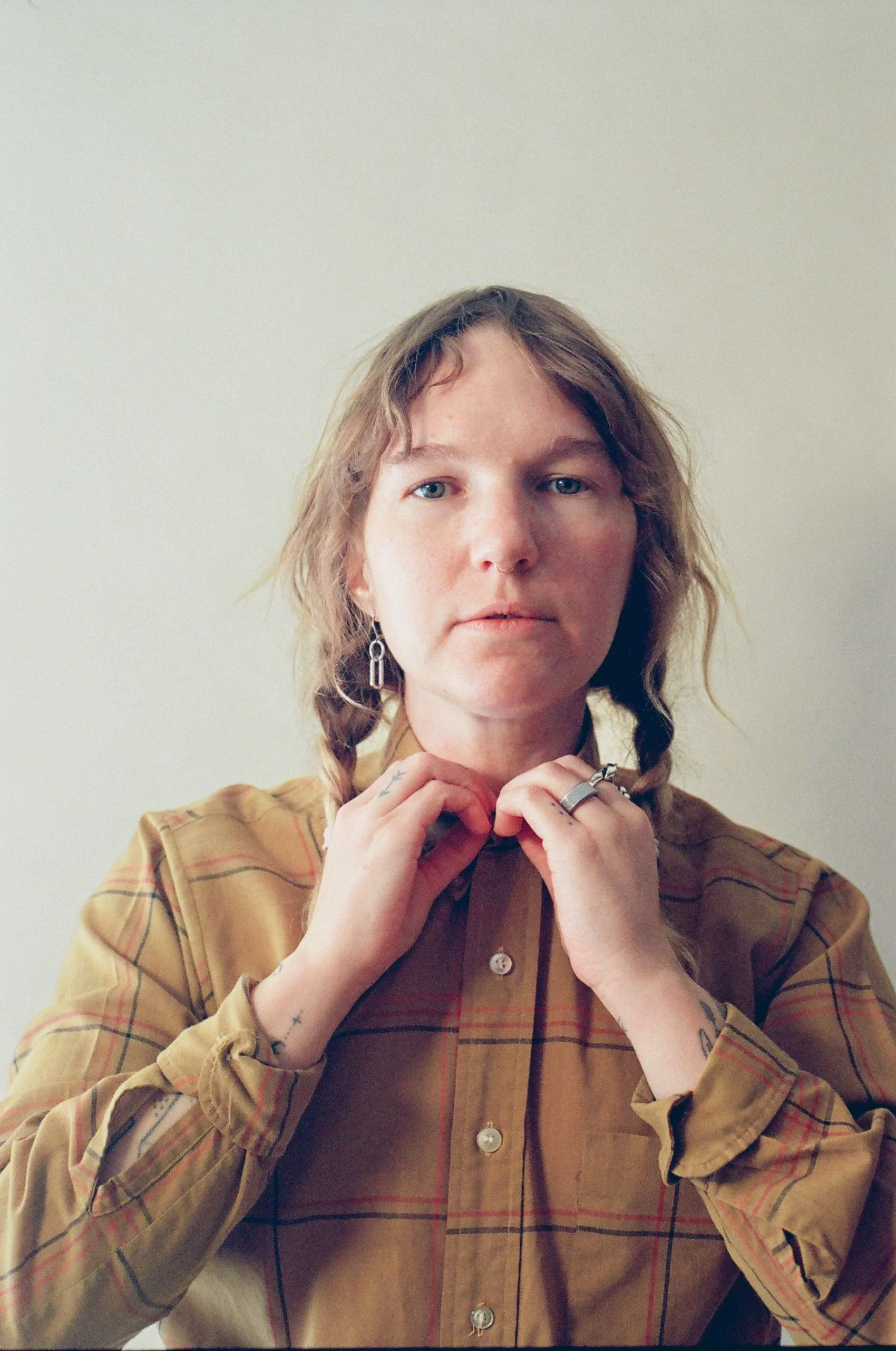 A person with braided hair, wearing a plaid button-up shirt, adjusting their collar, with rings on their fingers and earrings visible, standing against a plain, light-colored wall.