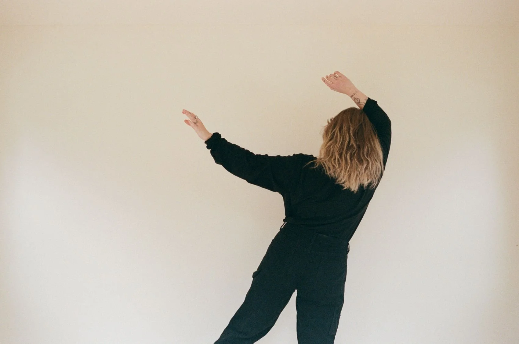 Person with wavy blonde hair, wearing black clothing, posing with arms raised and to the side against a plain off-white wall.