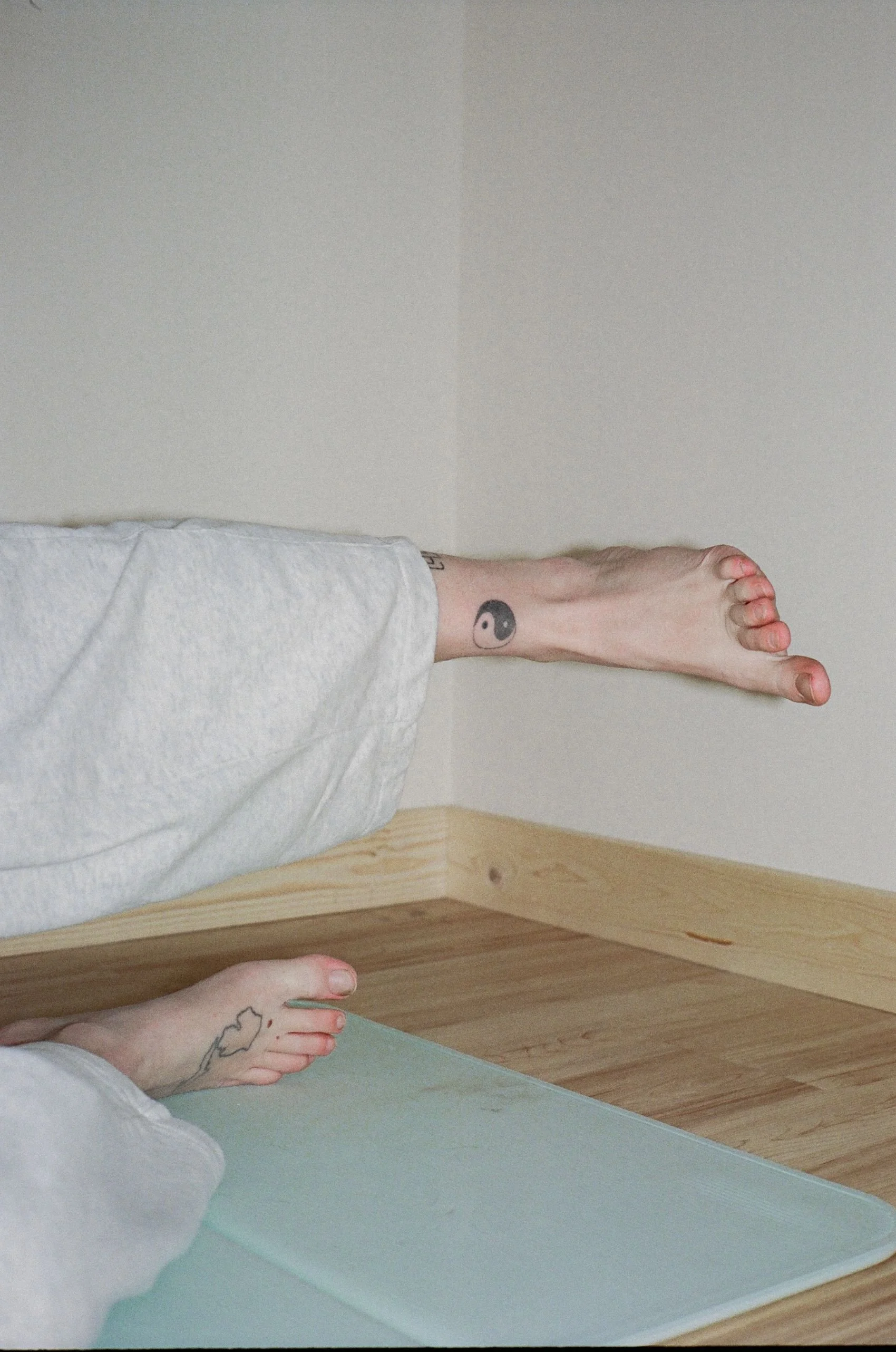 A person sitting on a yoga mat with one leg extended, showing tattoos on their foot and lower leg, near a corner of a room with wooden flooring and plain white walls.