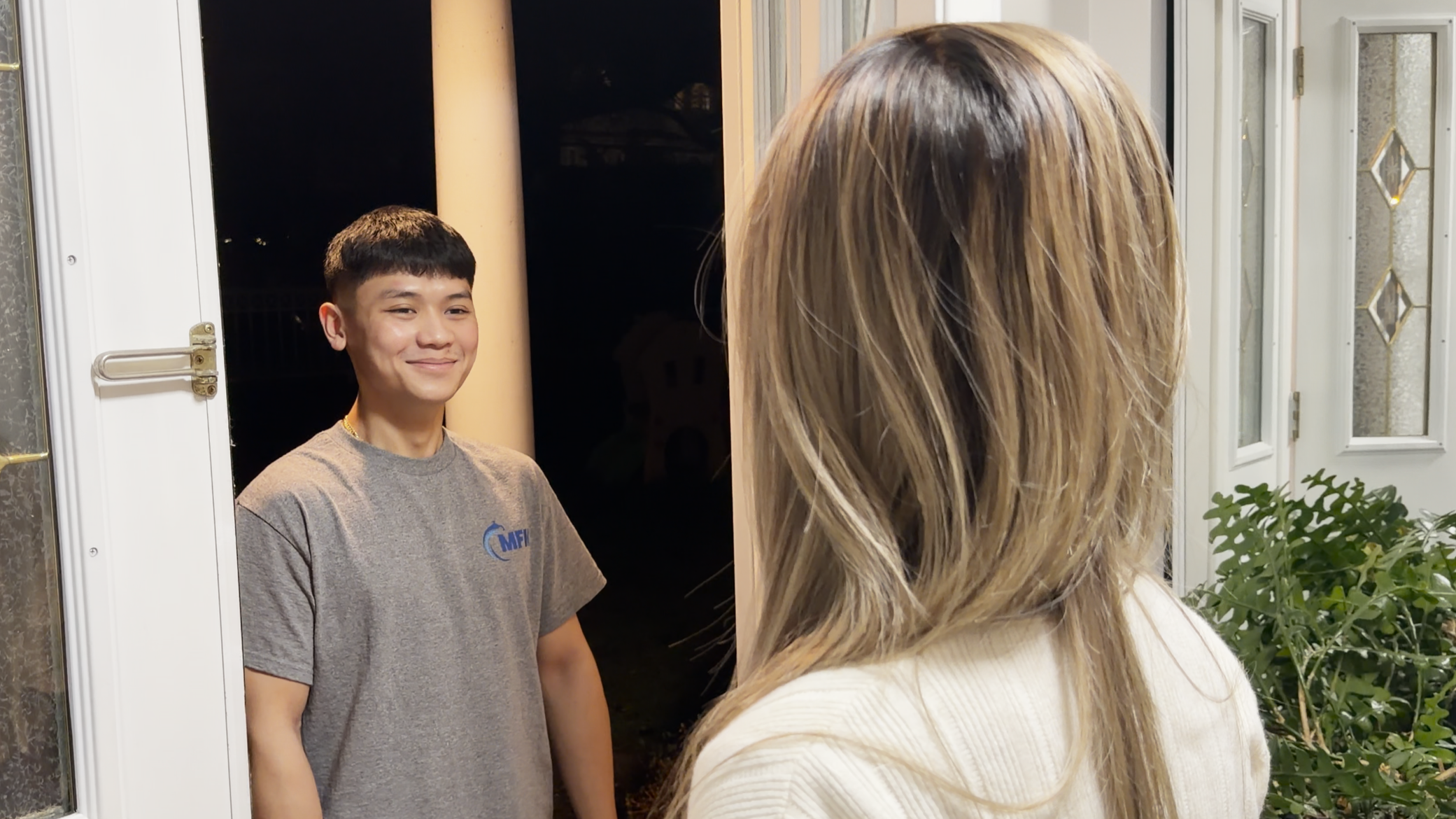 A young man in a gray t-shirt stands at a doorway at night, smiling as he greets a woman with long blonde hair, seen from behind, standing outside near a potted plant.