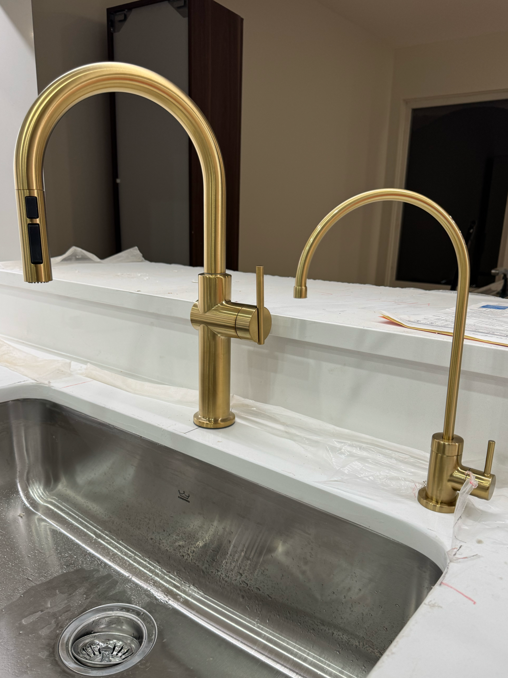 A kitchen countertop with two gold-colored modern kitchen faucets installed above a stainless steel sink, with a white paper or fabric covering the surface.
