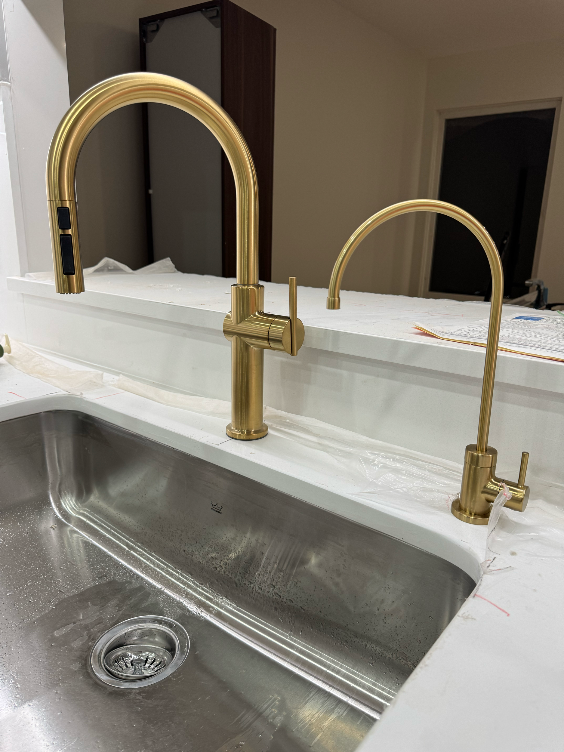 A kitchen sink with a gold faucet and a smaller gold side faucet, installed in a white countertop, with some plastic protective covering still on the surface.
