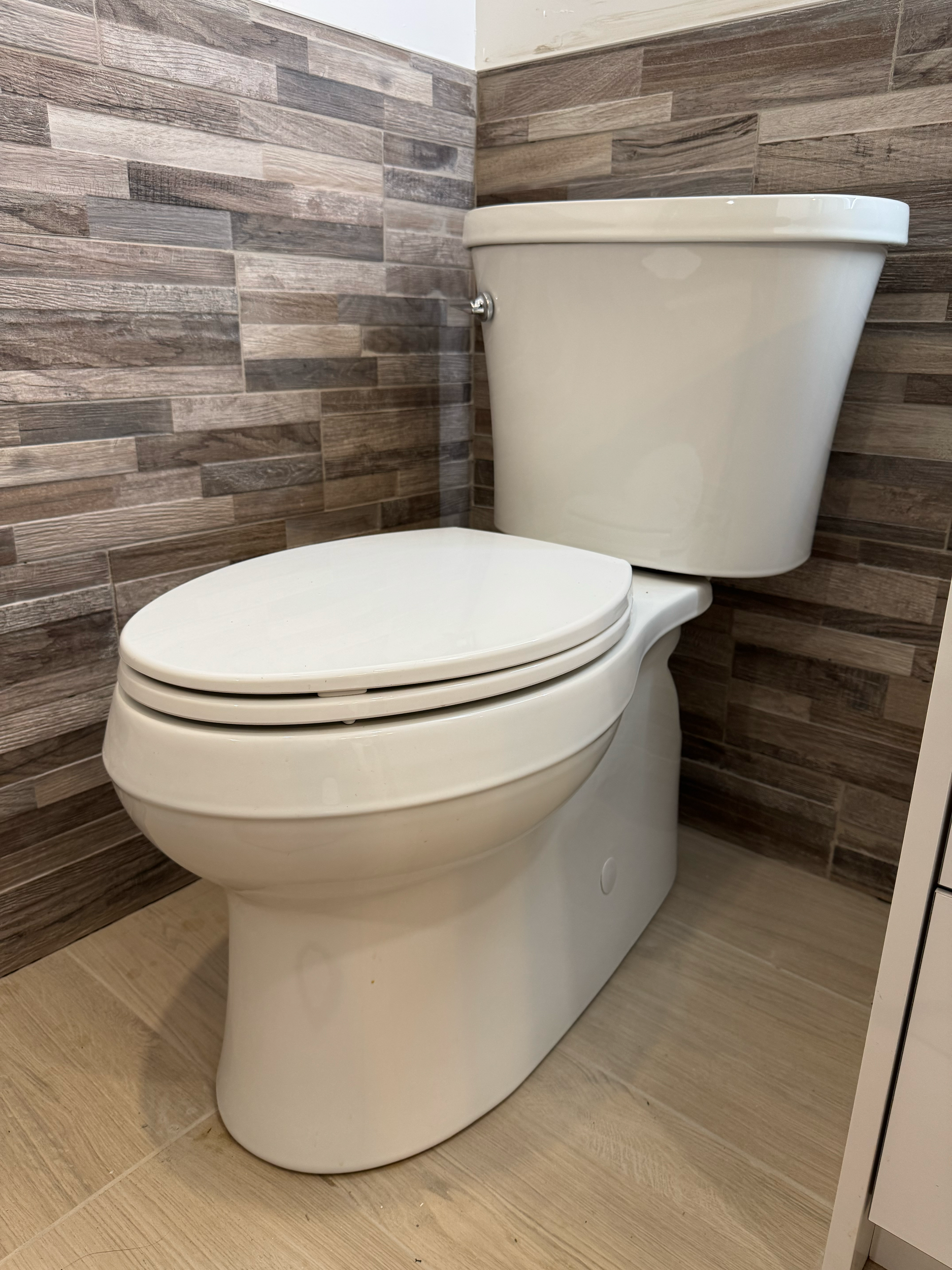 White ceramic toilet with a closed lid in a bathroom with wood-like wall tiles and light wood-colored floor tiles.