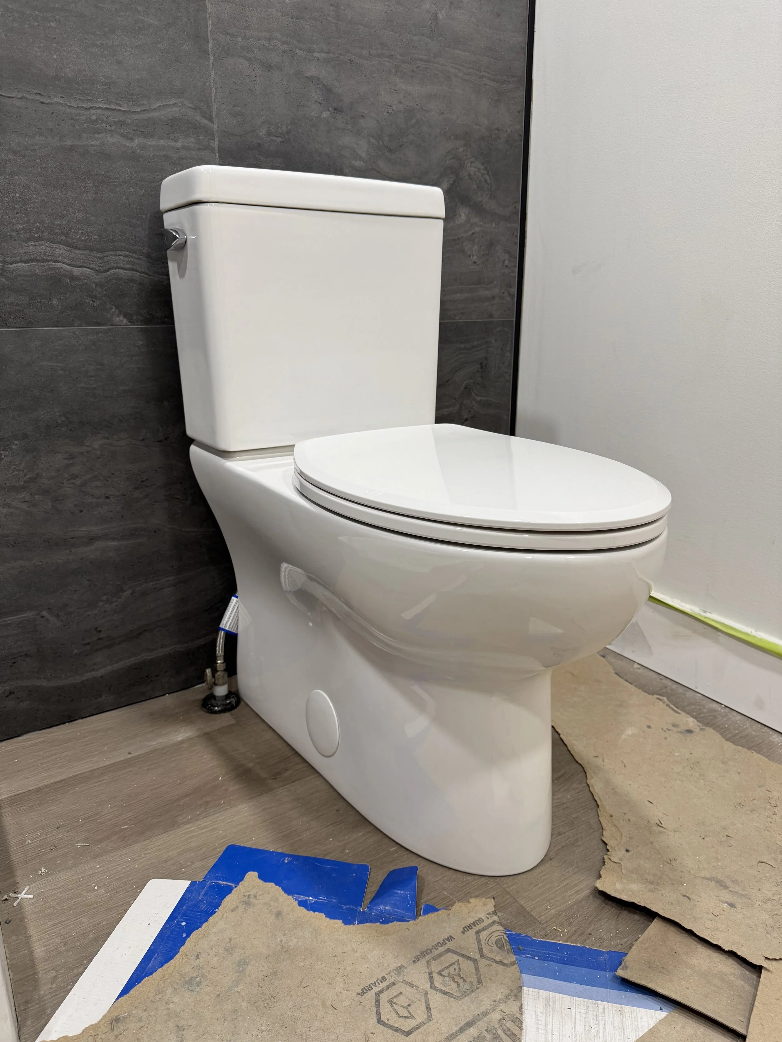 New white ceramic toilet installed against a dark gray tiled wall on a construction site with plywood and construction debris on the floor.