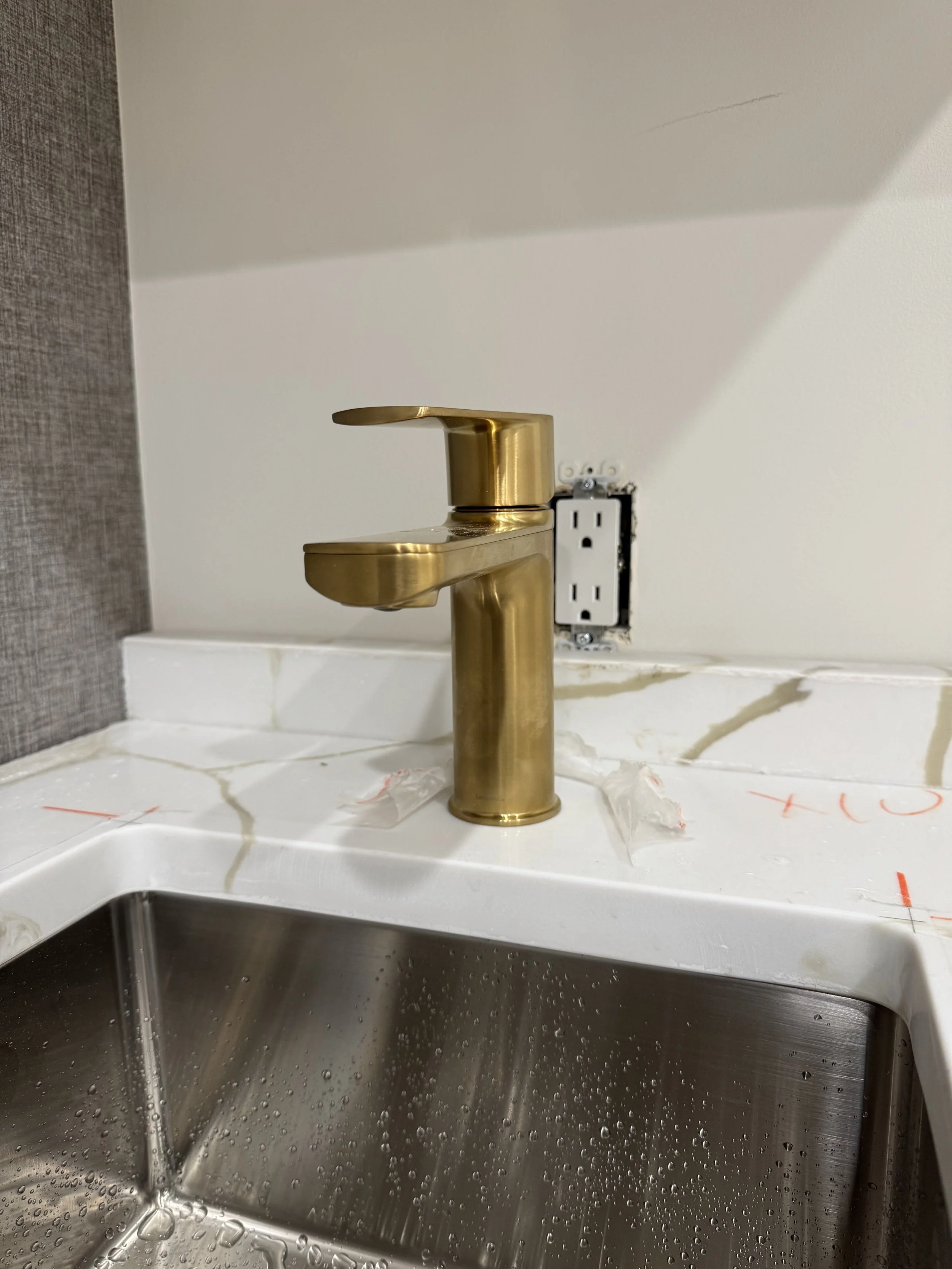 New gold-colored faucet installed above a stainless steel sink in a kitchen or laundry area. The surrounding countertop is white, and an electrical outlet is visible on the wall behind the faucet.