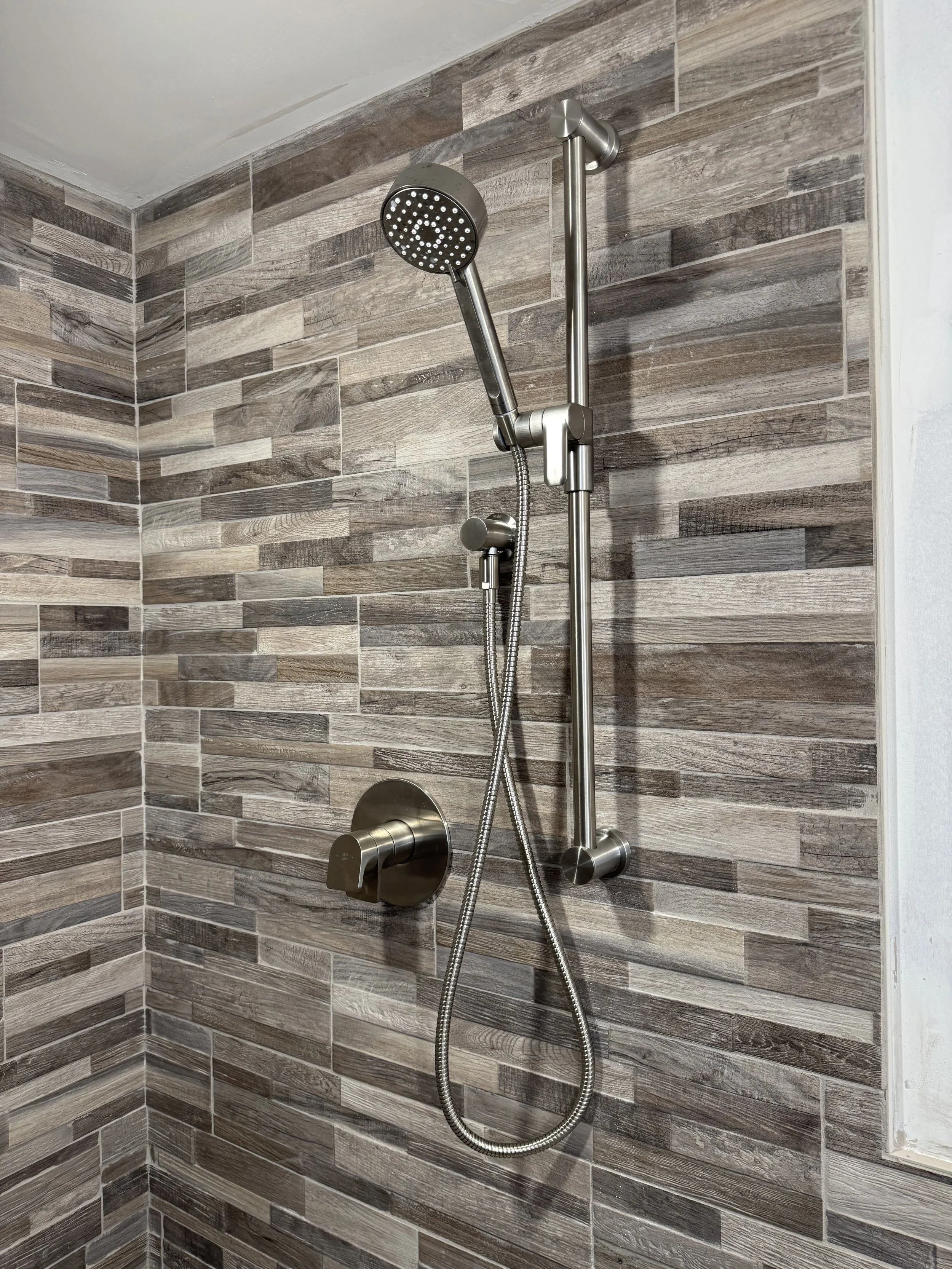 Stainless steel handheld showerhead and sliding bar mounted on a tiled wall in a bathroom shower.