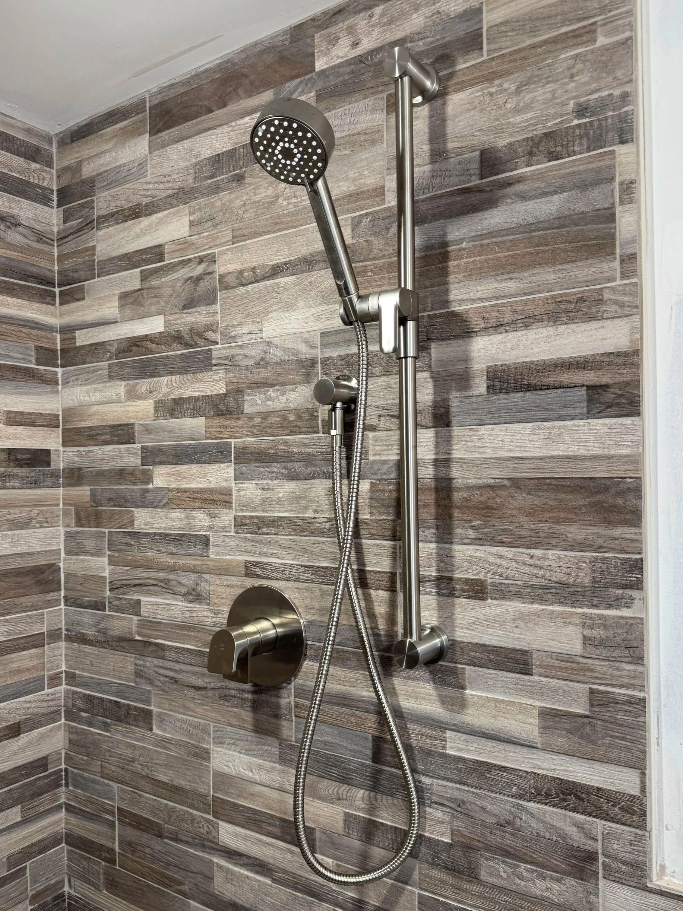Modern shower with a handheld and rain showerhead, mounted on a wood-textured tiled wall, featuring a wall-mounted handle and grab bar.