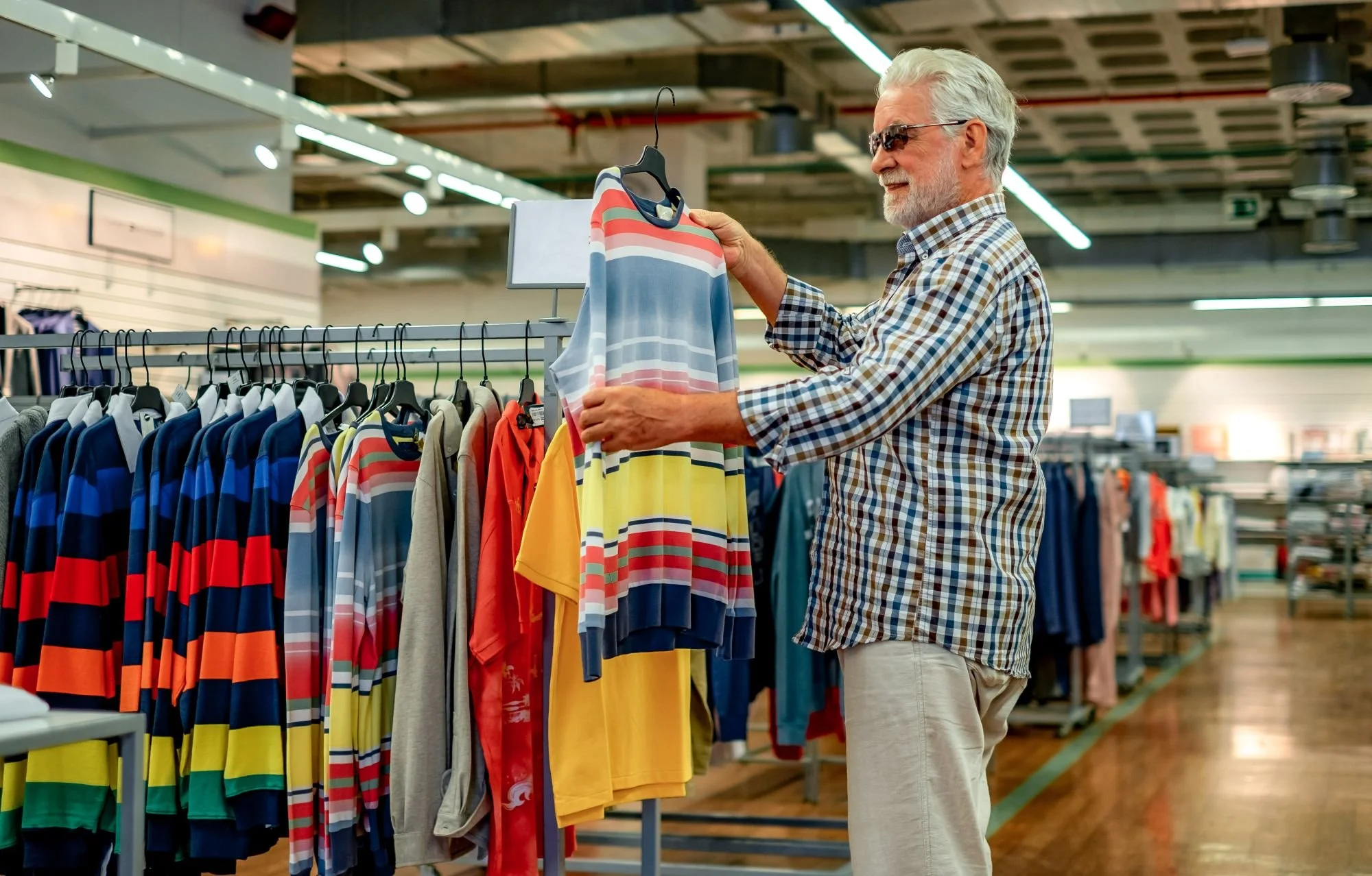 An elderly man with gray hair, wearing sunglasses, a checkered shirt, and beige pants, shopping for colorful striped shirts at a clothing store.