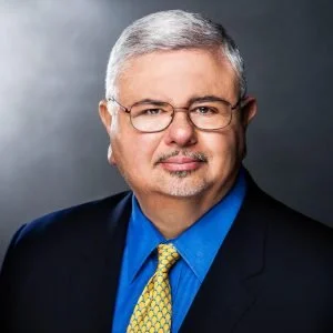 Portrait of a middle-aged man with gray hair, glasses, wearing a dark suit, blue shirt, and yellow tie, against a gray background.