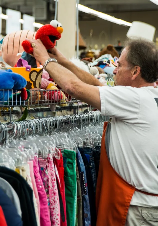 A man shopping for plush toys at a store, reaching for a red plush Elmo doll with white eyes and an orange nose.