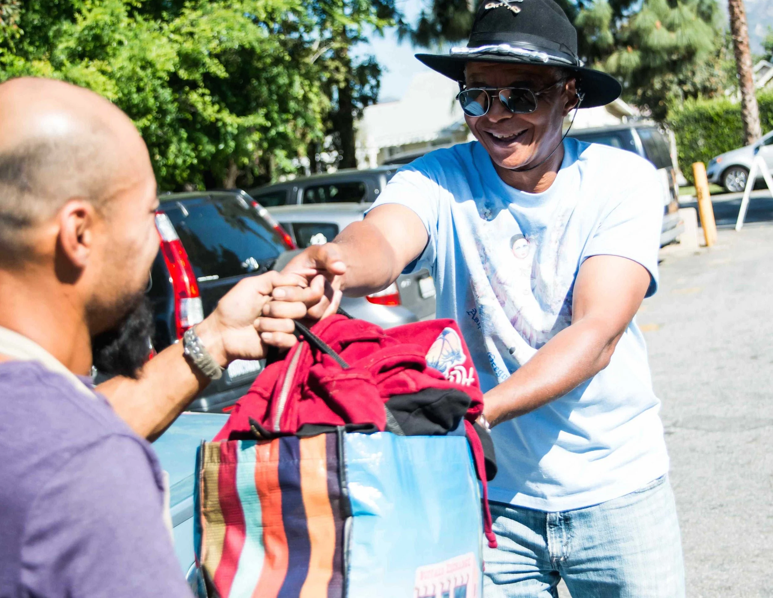 A woman in sunglasses and a wide-brimmed hat hands a red backpack to a man in a purple shirt in a parking lot with trees and parked cars in the background.
