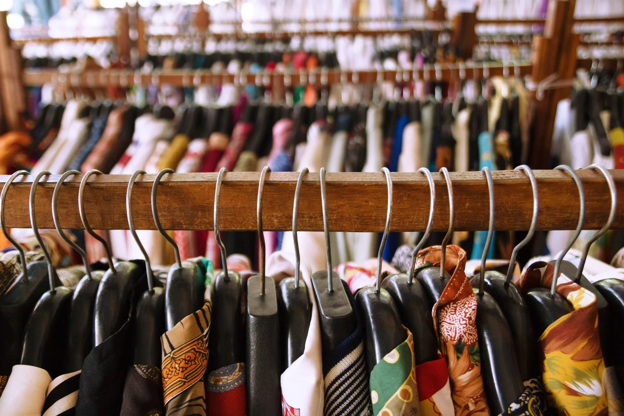 Clothes on hangers at a thrift store, with various colorful fabrics and patterns, in front of more clothing racks in the background.