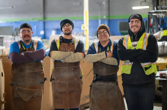 Four workers in safety vests and aprons standing with arms crossed in an industrial workshop.