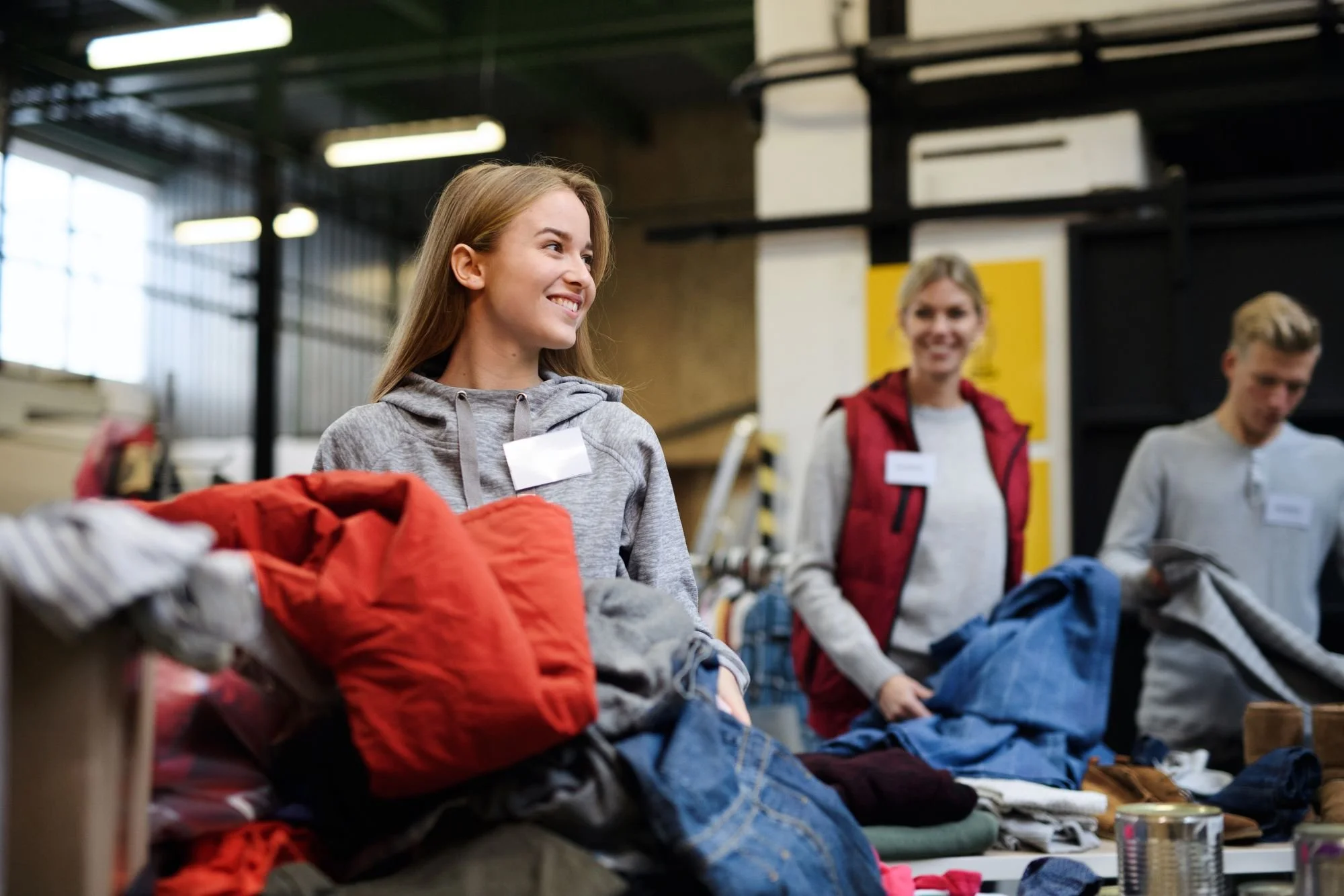 Three smiling people volunteering at a clothing drive, organizing clothes on tables in an industrial-style warehouse.