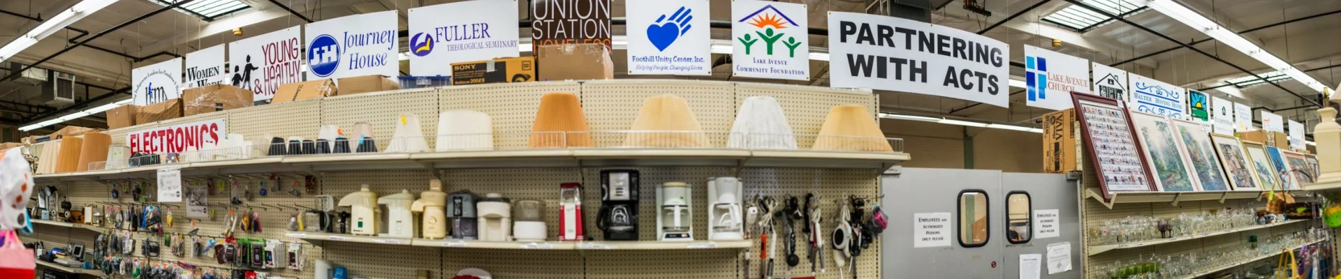 A retail store shelf displays assorted lamps, kitchen appliances, and electronic accessories. Above are various signs for community and church groups, with one reading 'Partnering with Acts.' The store has a ceiling with exposed beams and fluorescent lighting.