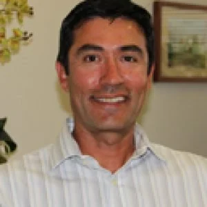 A man with short dark hair smiling, wearing a light-colored striped shirt, indoors with a painting and plant in the background.