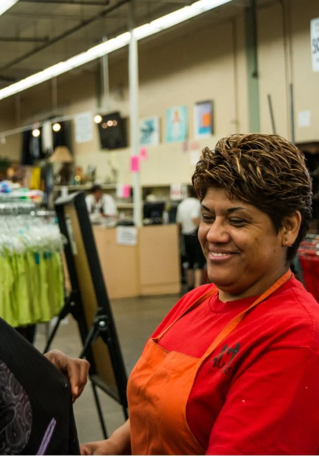 A woman with short curly hair wearing a red shirt and orange apron, smiling inside a store or restaurant.