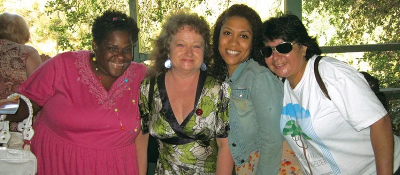 Four women smiling together outdoors, with a woman in pink, another in a floral dress, and two younger women, one with sunglasses.