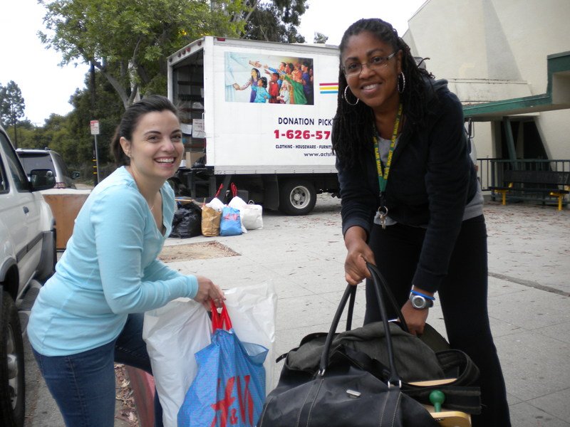 Two women smiling and holding bags outside near a donation truck, with various bags of donated items on the ground behind them.
