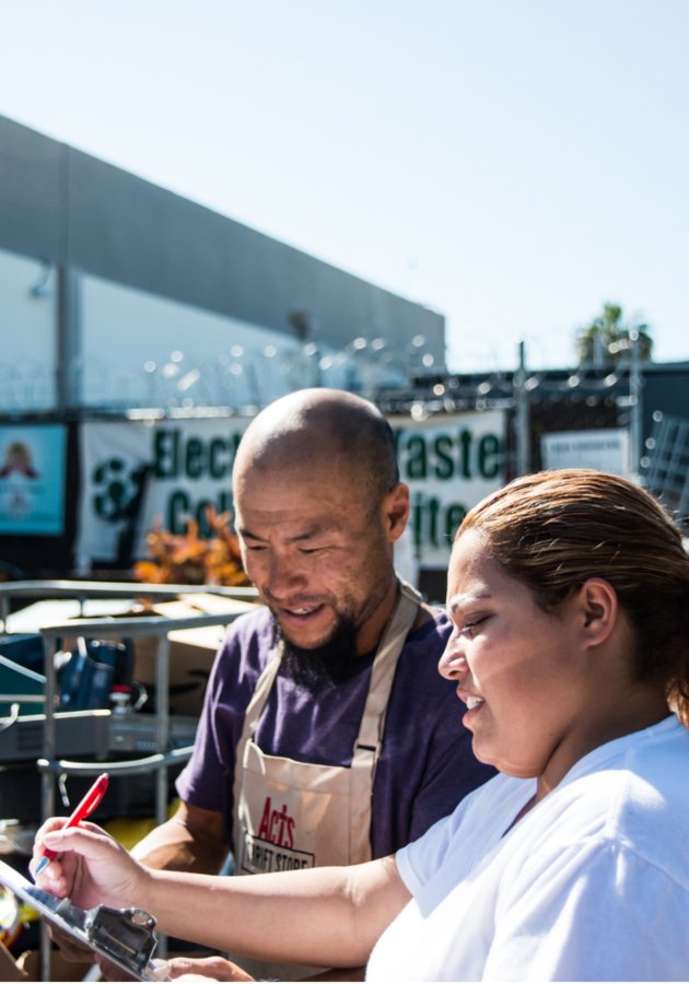 Two people, a man and a woman, outside at a flea market or outdoor event, looking at a clipboard or piece of paper together.