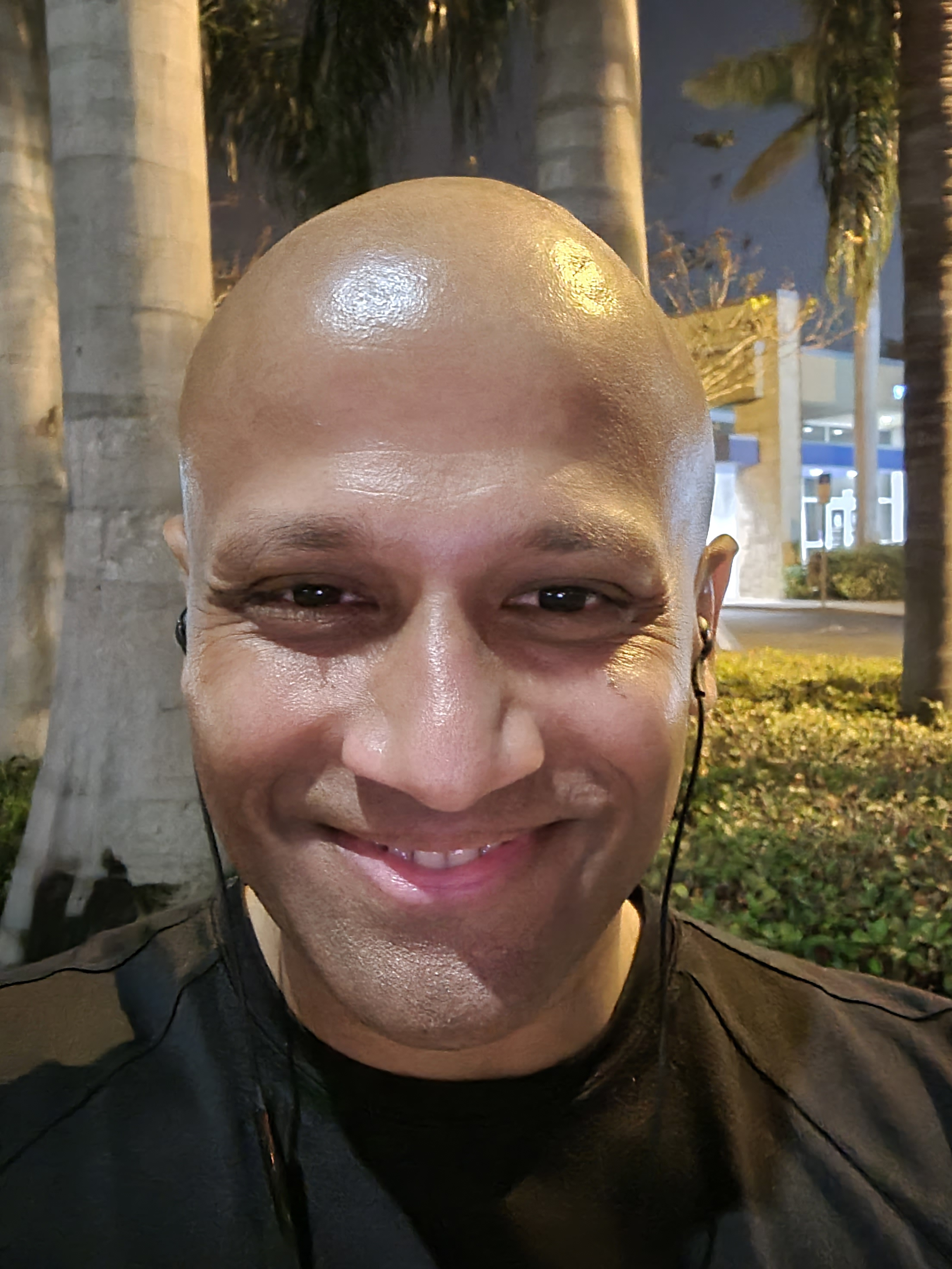 A smiling man with a shaved head and earphones, outdoors at night, with palm trees and a building in the background.