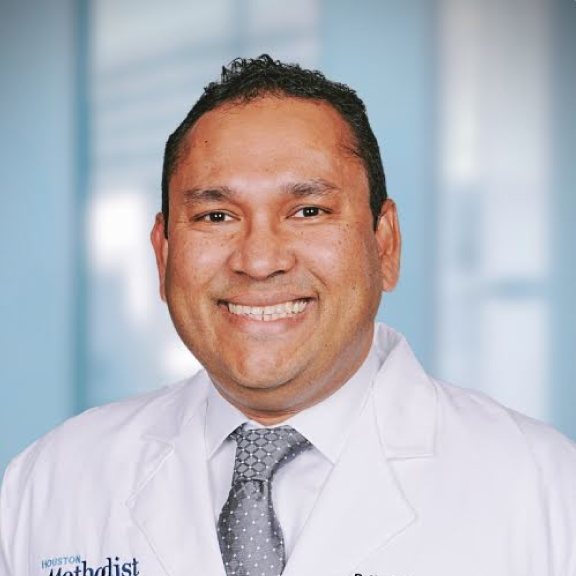 Smiling male doctor in white coat with 'Houston' embroidered on it, in front of a blurred office background.