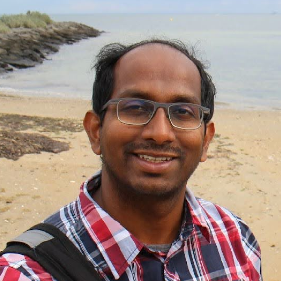 A man with glasses and a checkered shirt smiling on a beach with water and rocks in the background.