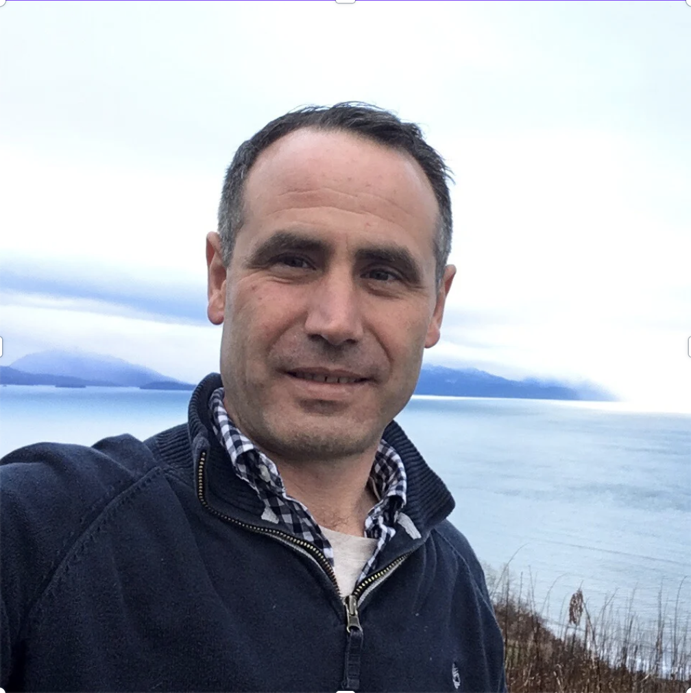 A man taking a selfie outdoors with a body of water, mountains, and cloudy sky in the background.