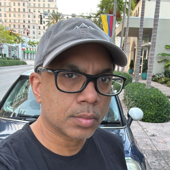 A man wearing a gray cap, black glasses, and a black shirt taking a selfie outdoors in an urban area with palm trees, colorful flags, and buildings in the background.