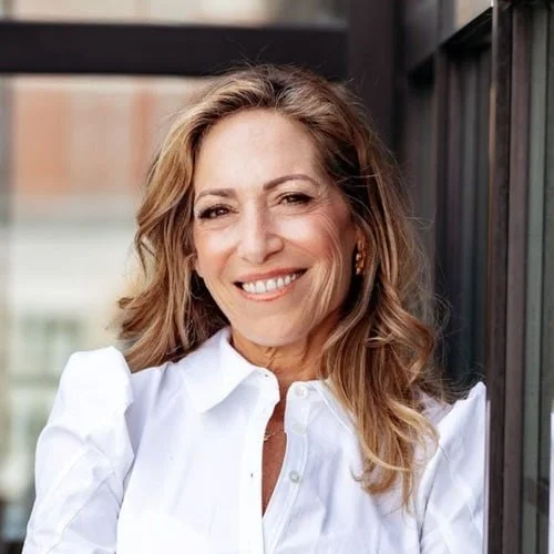 Smiling woman with wavy, light brown hair wearing a white shirt standing outdoors near a black fence.