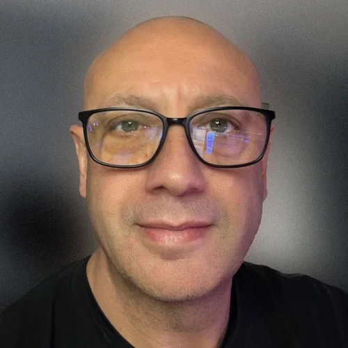 Close-up of a bald man wearing black-rimmed glasses and a black shirt, smiling gently against a dark background.