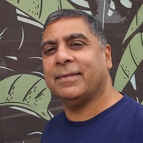 Close-up of a middle-aged man with short gray hair, wearing a dark blue T-shirt, standing in front of a decorative wall with green and brown leaves.
