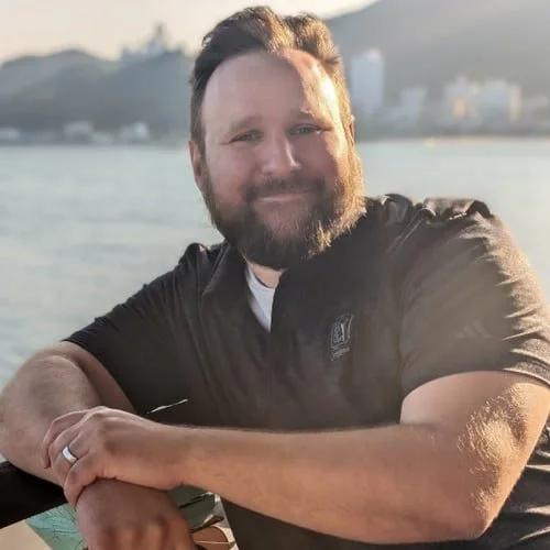 A man with a beard and short hair, smiling, wearing a black shirt, sitting outdoors near water with a hilly landscape and buildings in the background during sunset.