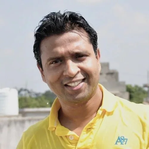 A smiling man with dark hair wearing a yellow polo shirt with an A7 logo, outdoors with a partly cloudy sky in the background.