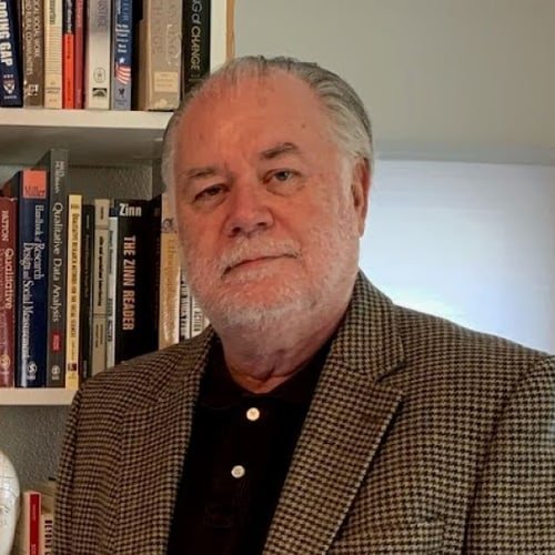Portrait of an older man with gray hair and a beard, wearing a checkered blazer and black shirt, standing in front of a bookshelf filled with books.