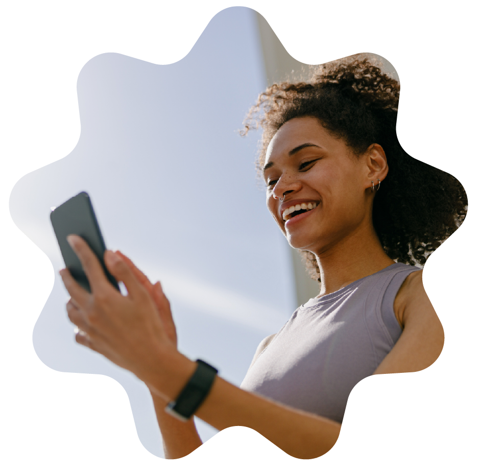 Smiling woman with curly hair, earrings, and a nose ring looking at her smartphone outside on a sunny day.