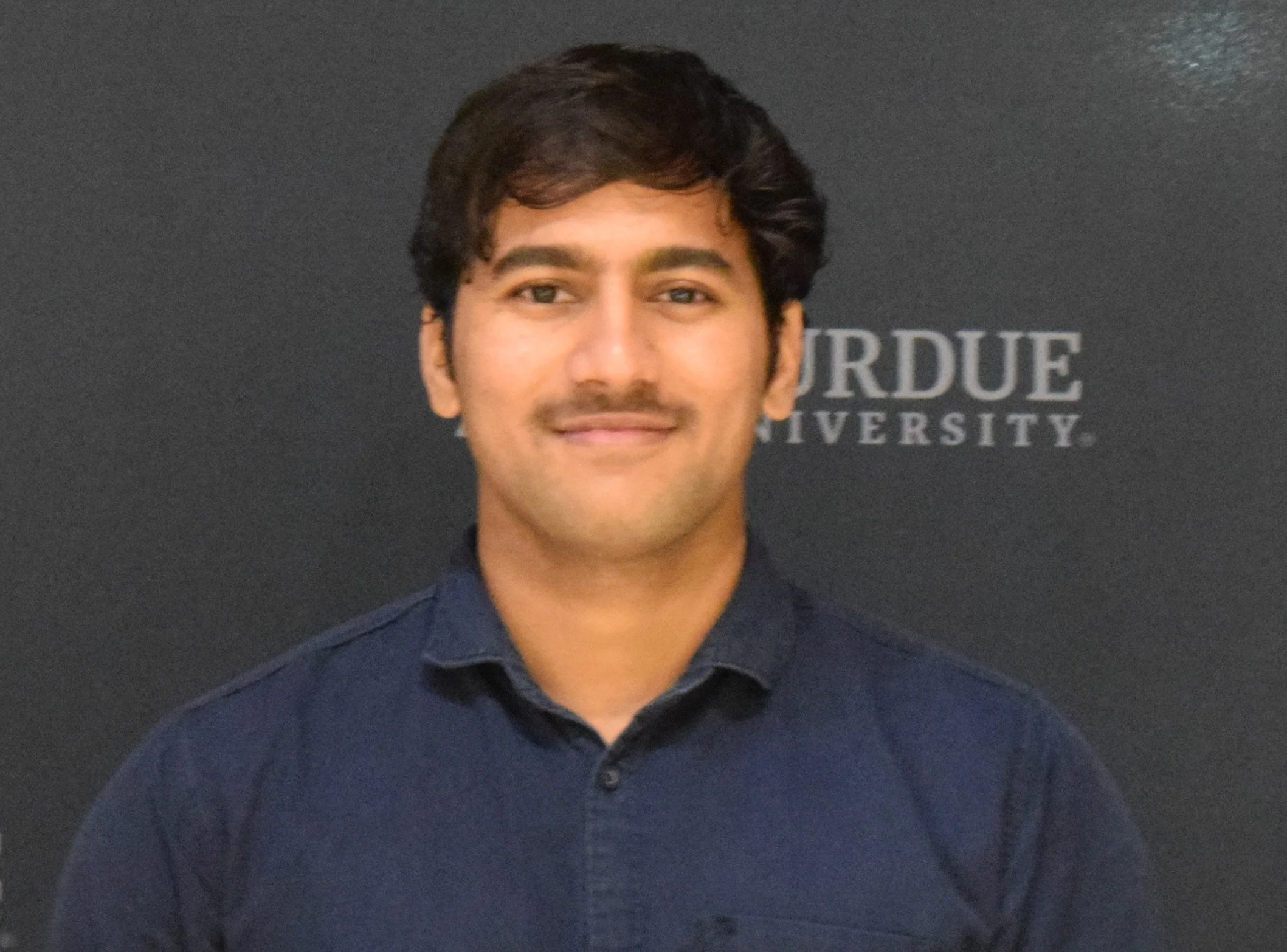 Young man with dark hair and a slight smile, wearing a dark shirt, standing in front of a black background with the word "PURDUE" and "UNIVERSITY" partially visible.