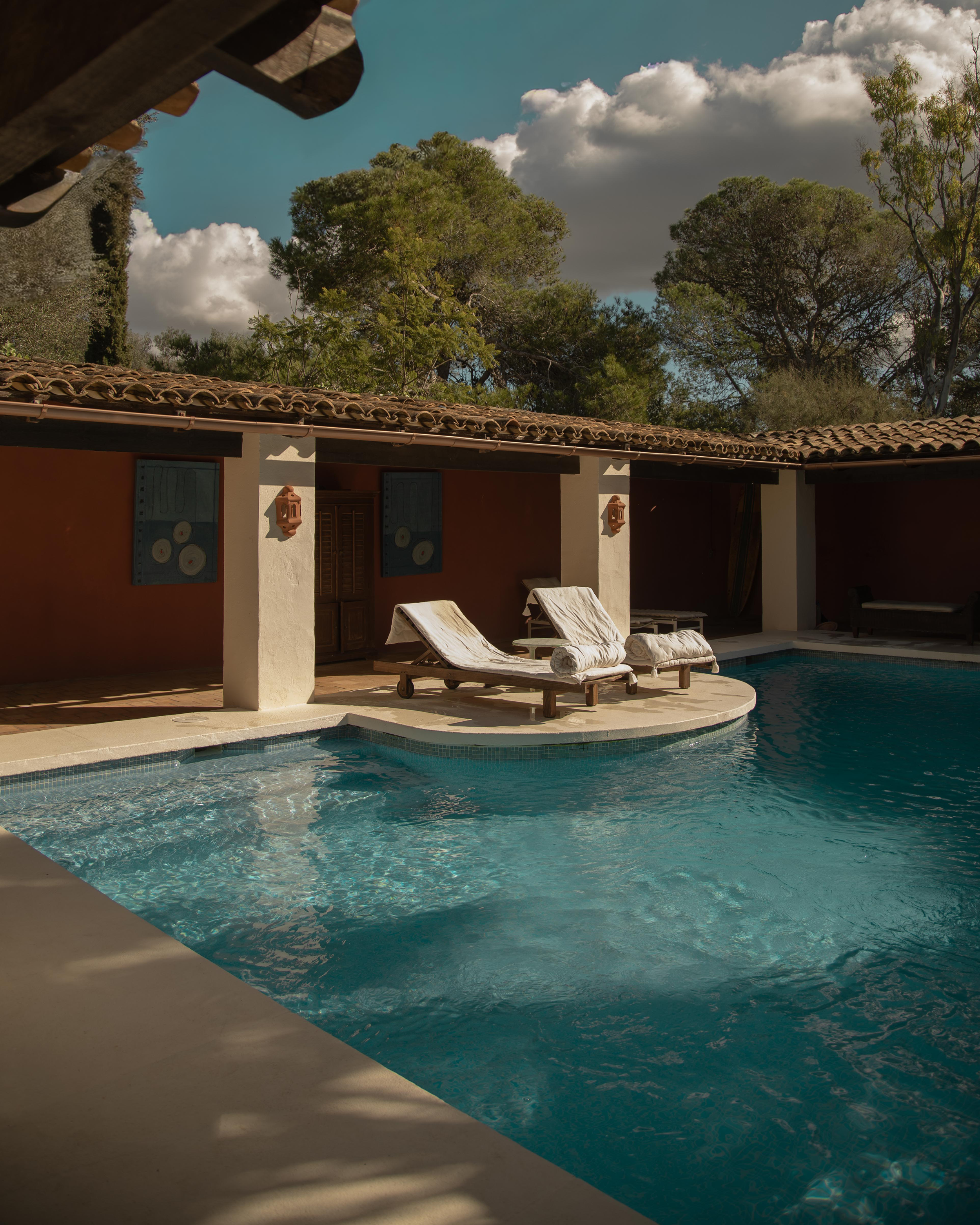 A peaceful outdoor pool area at a villa or resort with lounge chairs, rolled towels, a shaded structure, and trees in the background.