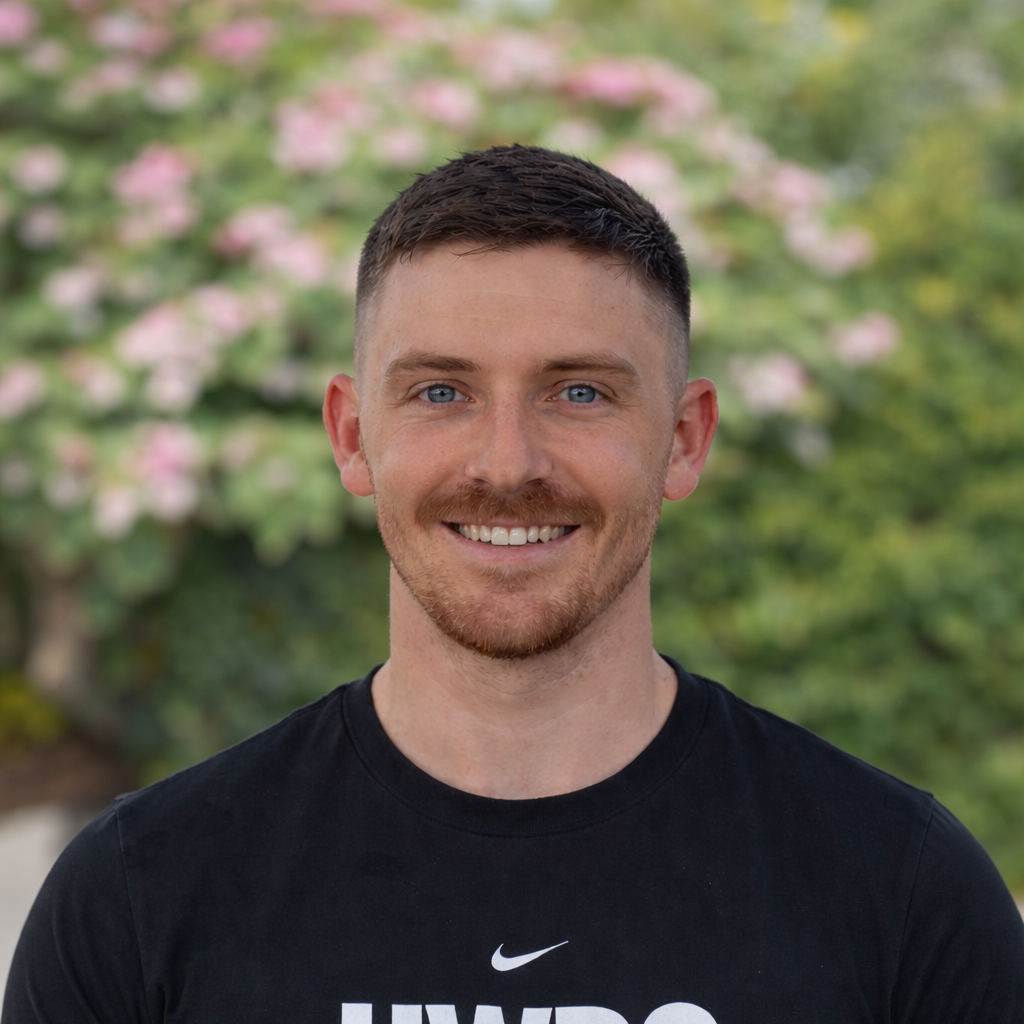 A young man with short brown hair and blue eyes, smiling, wearing a black Nike t-shirt, outdoors with blurred green trees and pink flowers in the background.