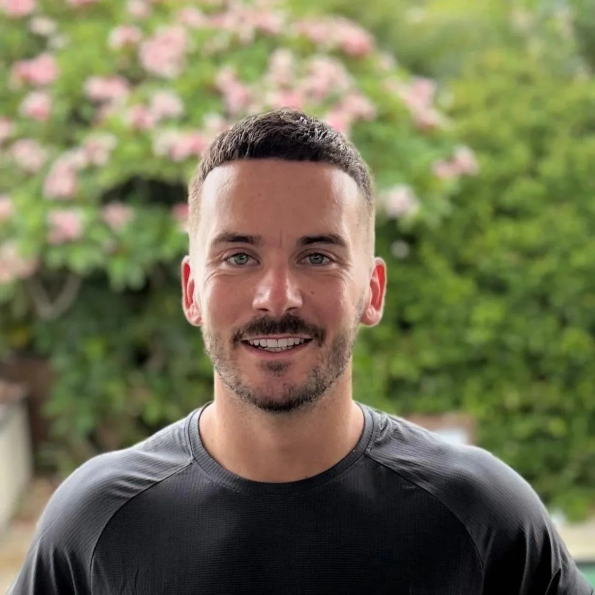A young man with short dark hair, a beard, and blue eyes, smiling at the camera. He is wearing a black athletic shirt, standing outdoors with green trees and pink flowers in the background.