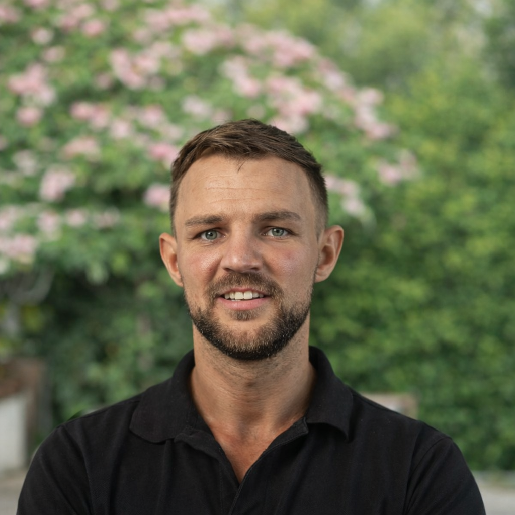 A man with short brown hair and blue eyes, wearing a black collared shirt, smiling outdoors with trees and pink flowers in the background.